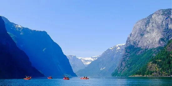 Group of kayakers paddling on a wide, calm body of water surrounded by tall, rugged mountains with patches of snow under a clear blue sky.