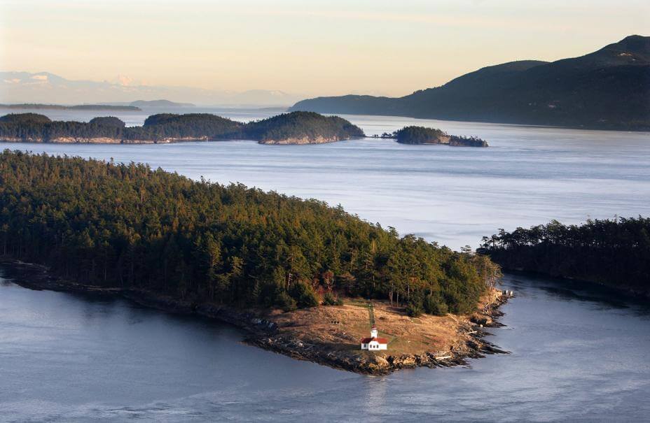 Aerial view of a small tree covered island with a white house near the shore surrounded by calm waters and additional islands in the background