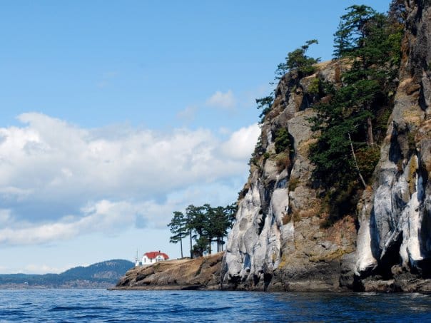 image.jpeg | Sea Quest Kayak Tours A scenic view of a rocky cliff with trees and a small white house with a red roof at its base set against a backdrop of blue sky and distant mountains