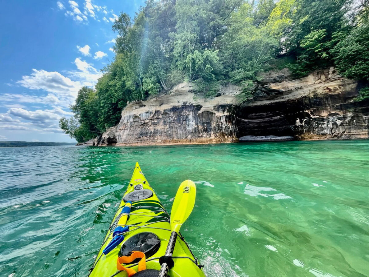Guided Kayak Tours in the San Juan Islands 2 Experience the view from a yellow kayak on clear turquoise water, approaching rocky cliffs and caves with green trees overhead under a partly cloudy sky—perfect for those seeking unforgettable Guided Kayak Tours.