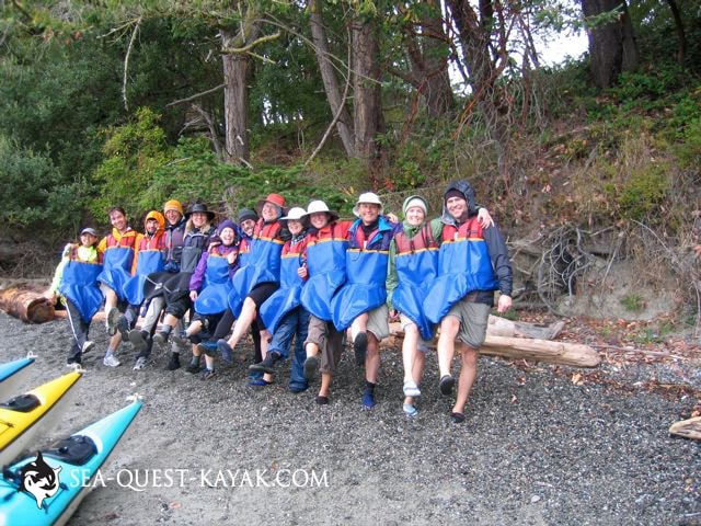 A group of people in kayaking gear from Kayak-Rentals-San-Juan-Islands sit on a log by the shore with their legs raised, posing for a group photo; kayaks and trees are visible nearby.