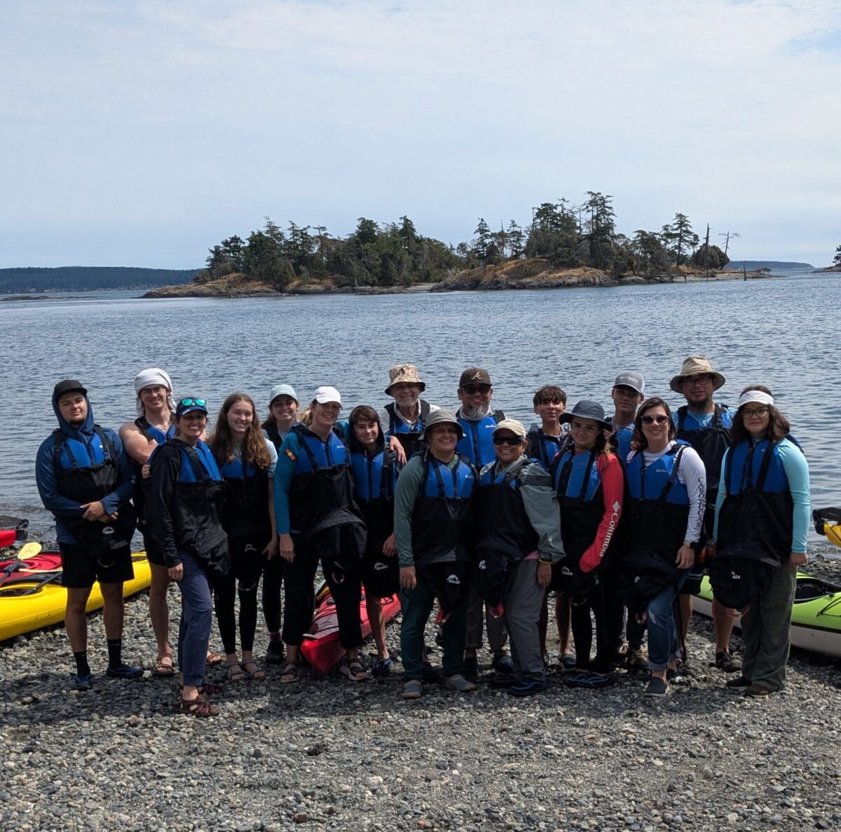 A group of people in life jackets stand on a rocky shore in front of kayaks, ready for a Kayak Tour Seattle adventure, with a body of water and small forested islands in the background.