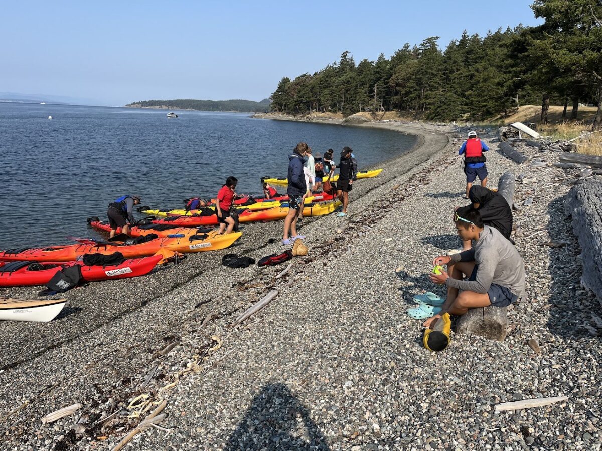A group of people prepare kayaks on a rocky beach by the water, with trees and kayaks lining the shoreline—capturing the spirit of kayaking Seattle’s scenic waterways.