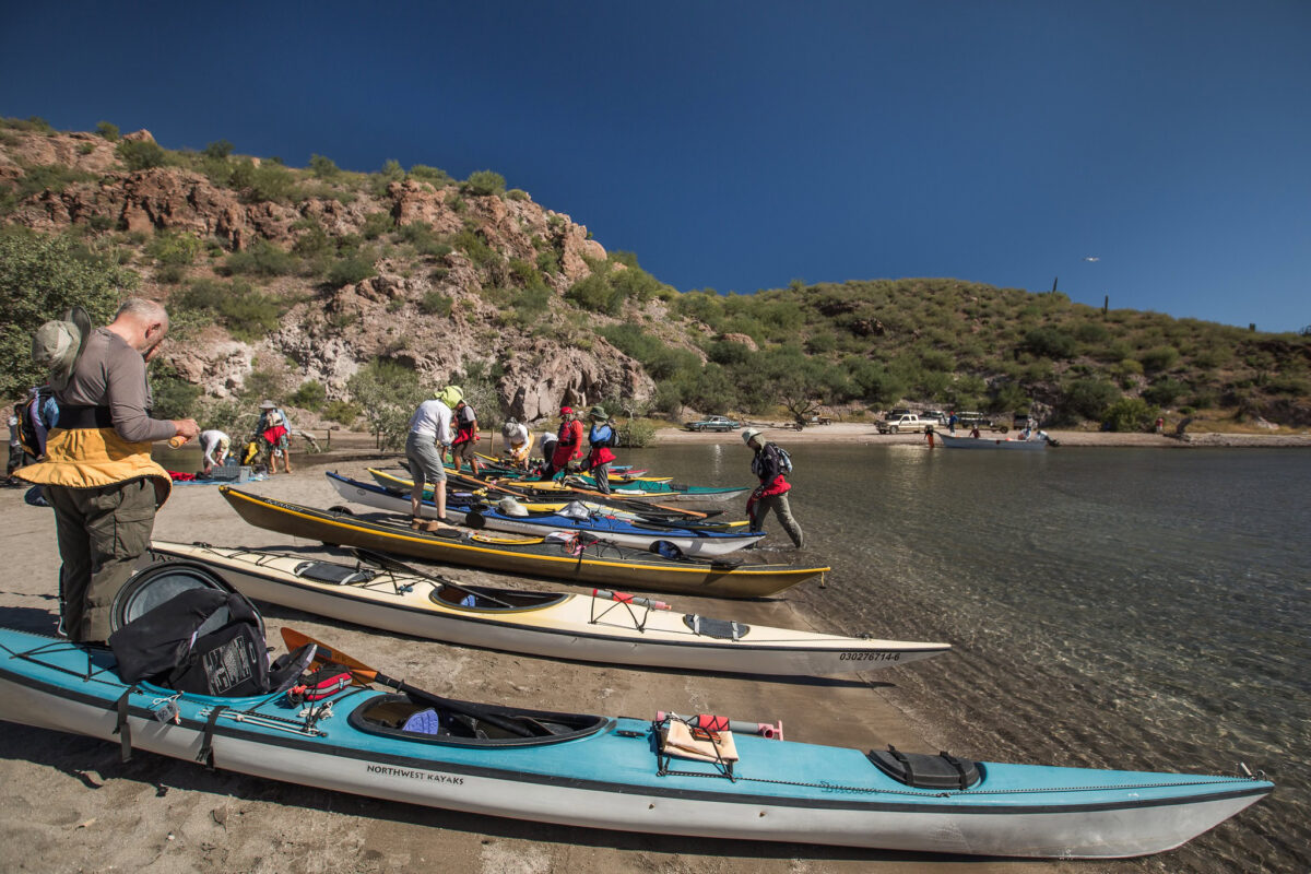 People prepare kayaks on a sandy shore near clear water, with rocky hills and vegetation in the background under a clear blue sky—perfect for San Juan Island Kayaking Trips.