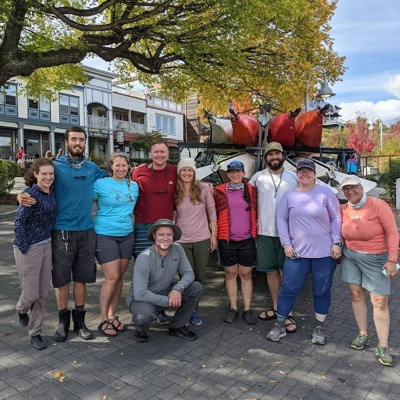 A group of ten people pose smiling under a tree with kayaks stacked on a trailer behind them, enjoying a sunny day in an urban outdoor setting—perfect for those seeking San Juan Islands Lodging and adventure.