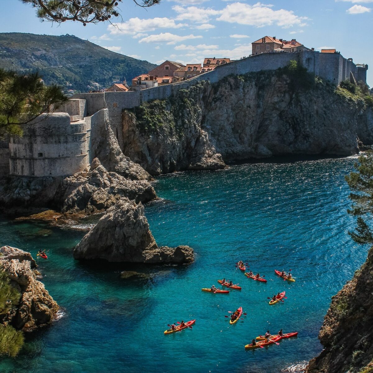 A group of kayakers paddles in clear blue water below rocky cliffs and historic stone buildings under a partly cloudy sky, just moments away from charming San Juan Islands lodging.