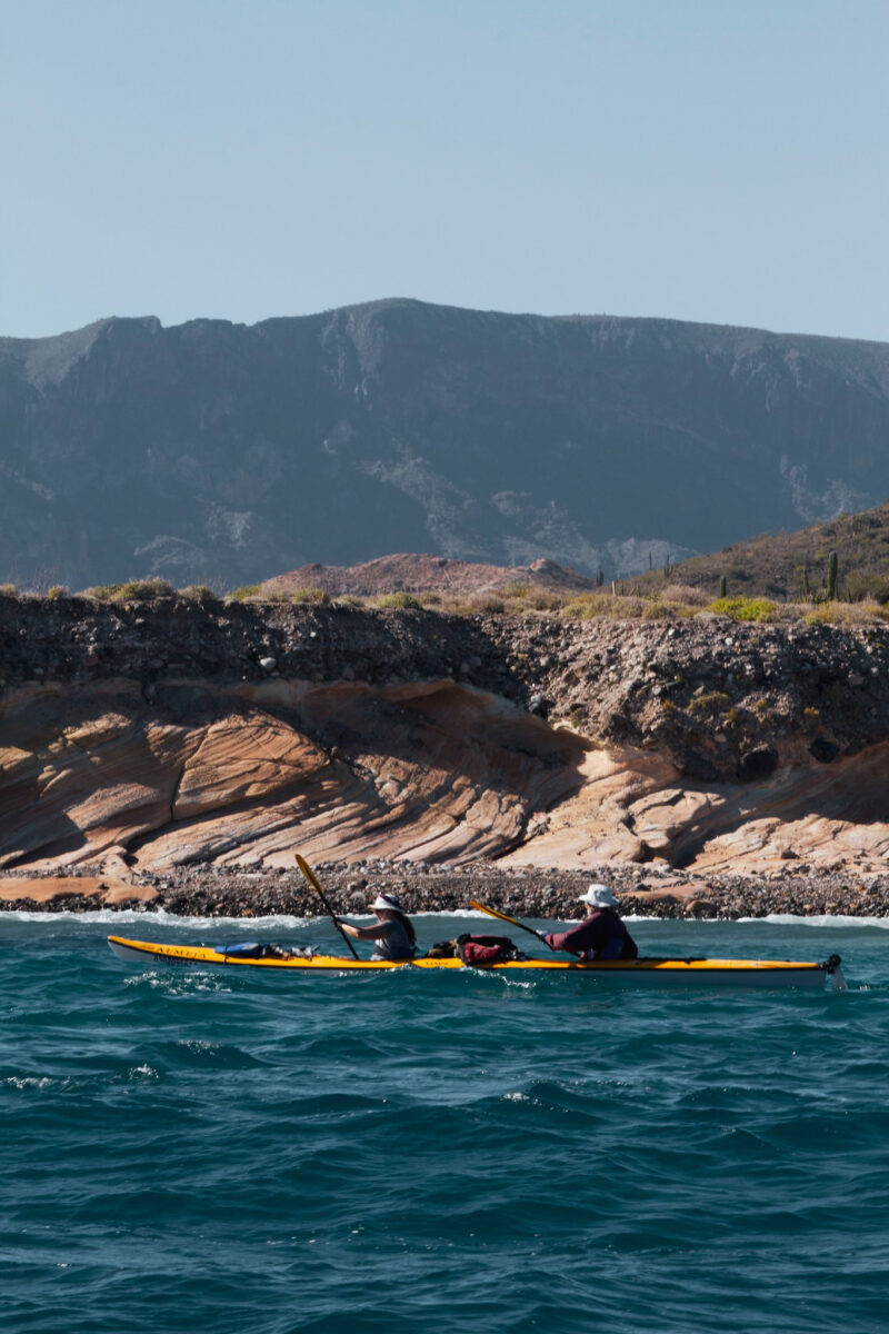 Two people enjoy a Kayak Tour, paddling a yellow kayak on blue water near a rocky shoreline with mountains in the background.