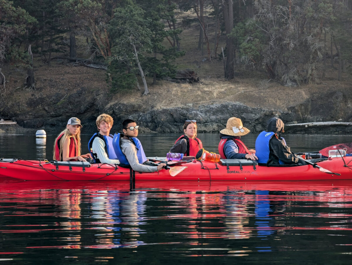 A group of six people wearing life jackets sit in a red kayak on calm water near a wooded shoreline, enjoying kayaking Seattle style.