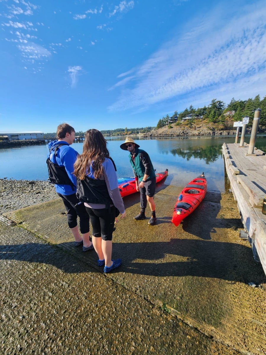 Three people stand on a boat ramp near two red kayaks at Friday Harbor, listening to an instructor by the water under a blue sky—perfect for a day of kayaking.