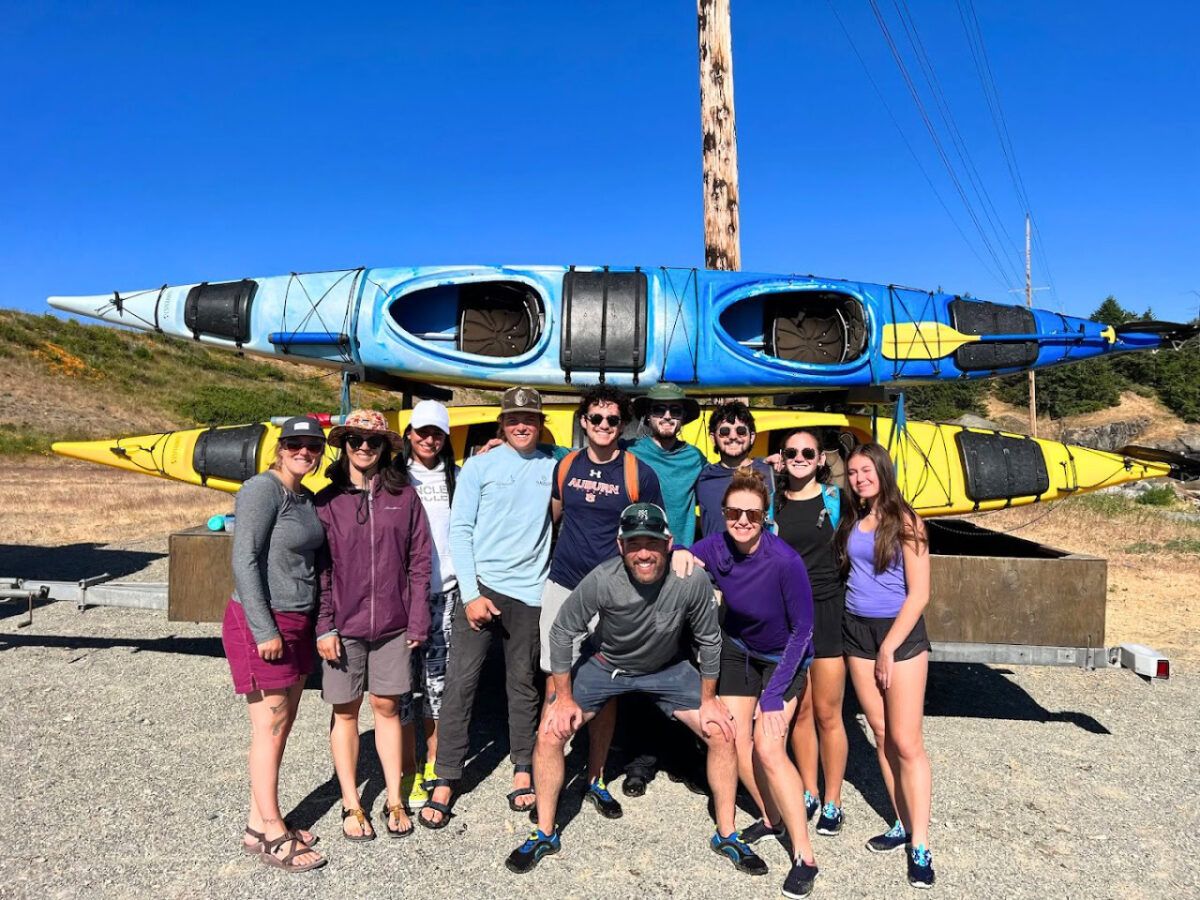 A group of ten people pose and smile in front of stacked blue and yellow kayaks outdoors on a sunny day, ready to embark on one of the unforgettable San Juan Island Kayaking Trips.