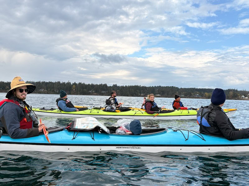 Guided Kayak Tours in the San Juan Islands 1 Five people in two kayaks paddle on a calm body of water during one of our Guided Kayak Tours, with a forested shoreline and cloudy sky in the background. Maps and gear are visible on the front kayak.