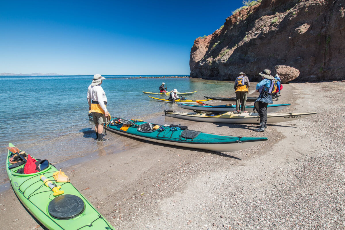 Several people prepare kayaks on a sandy beach beside clear blue water and rocky cliffs under a bright sky, ready to embark on unforgettable San Juan Island Kayaking Trips.