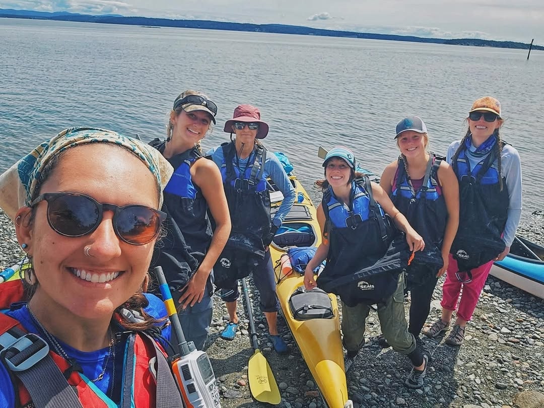Six people in life vests pose and smile near kayaks on a rocky shore with calm water, enjoying their Kayak-Rentals-San-Juan-Islands adventure with distant land in the background.