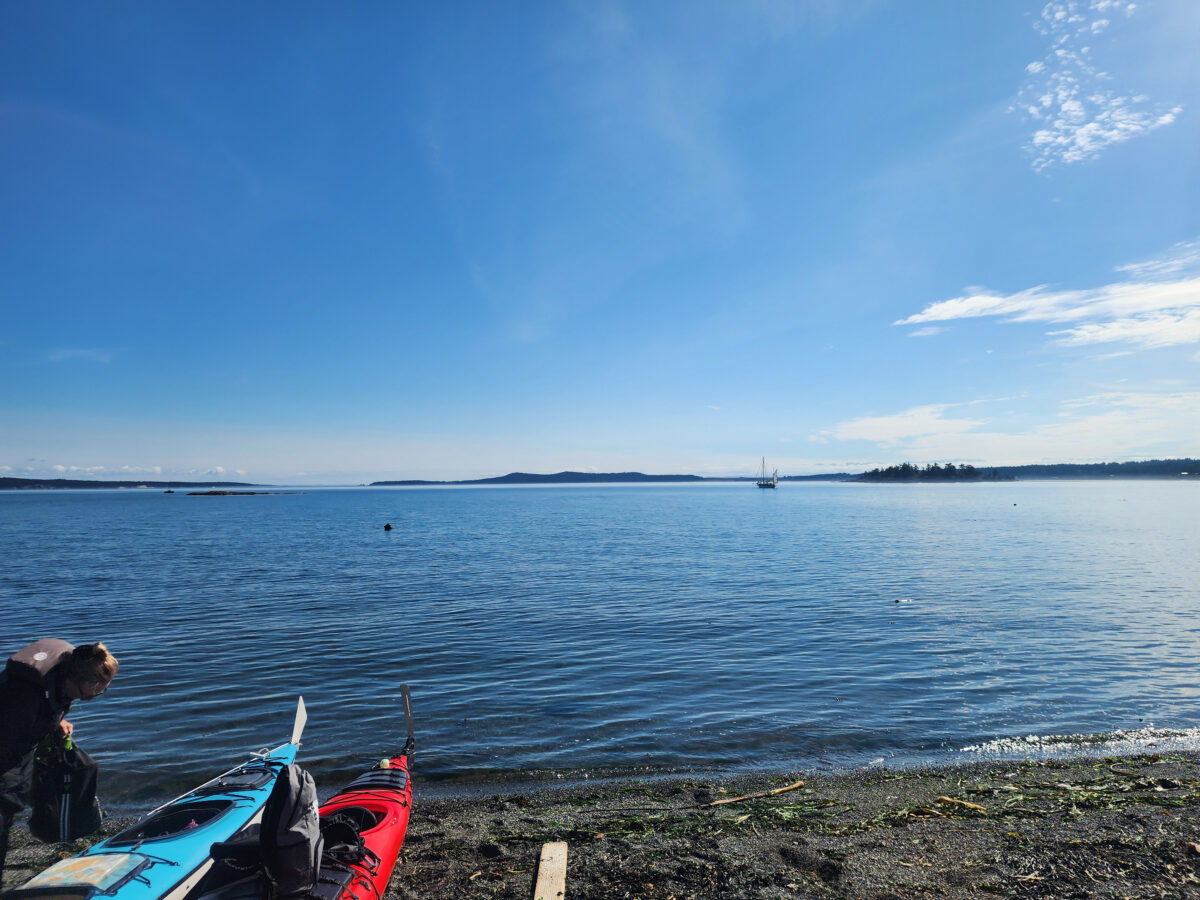 A calm beach scene with two kayaks on the shore, a person bending near them, and serene blue water stretching toward distant islands—perfect for those dreaming of San Juan Islands Boat Tours under a clear sky.