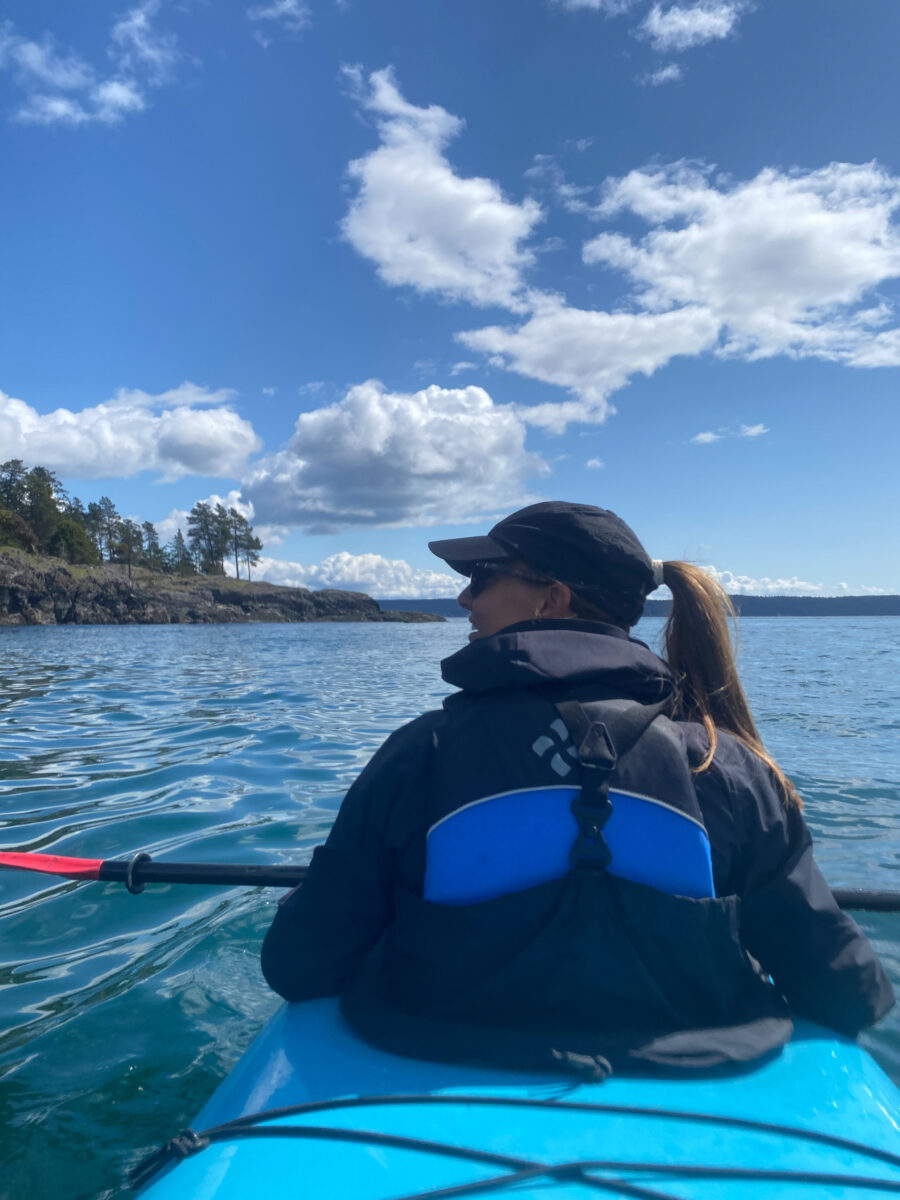 A person kayaking on a blue lake near a rocky shoreline under a partly cloudy sky, facing away from the camera—an adventure inspired by San Juan Islands Boat Tours.