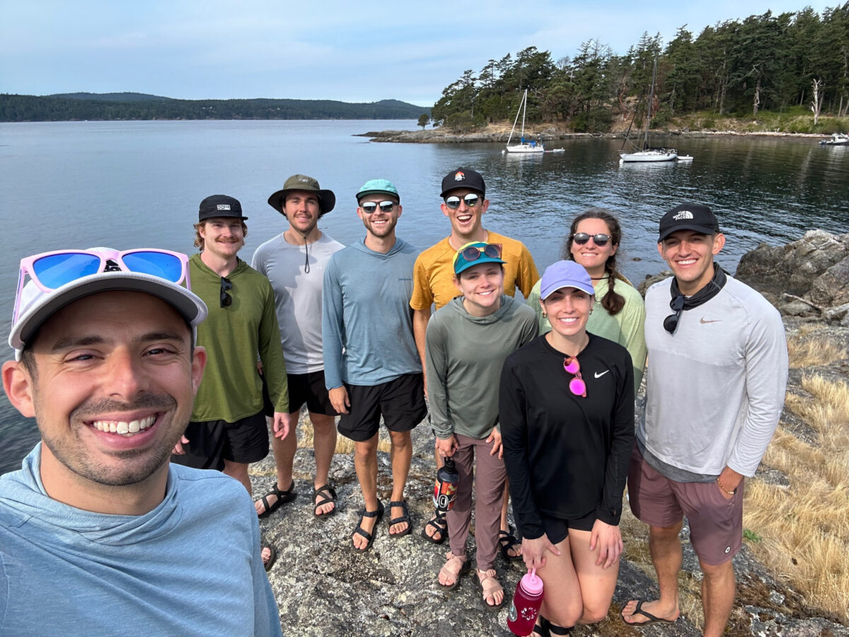 A group of nine people poses on a rocky shoreline with water, boats, and trees in the background on a cloudy day during their San Juan Islands Boat Tours adventure.