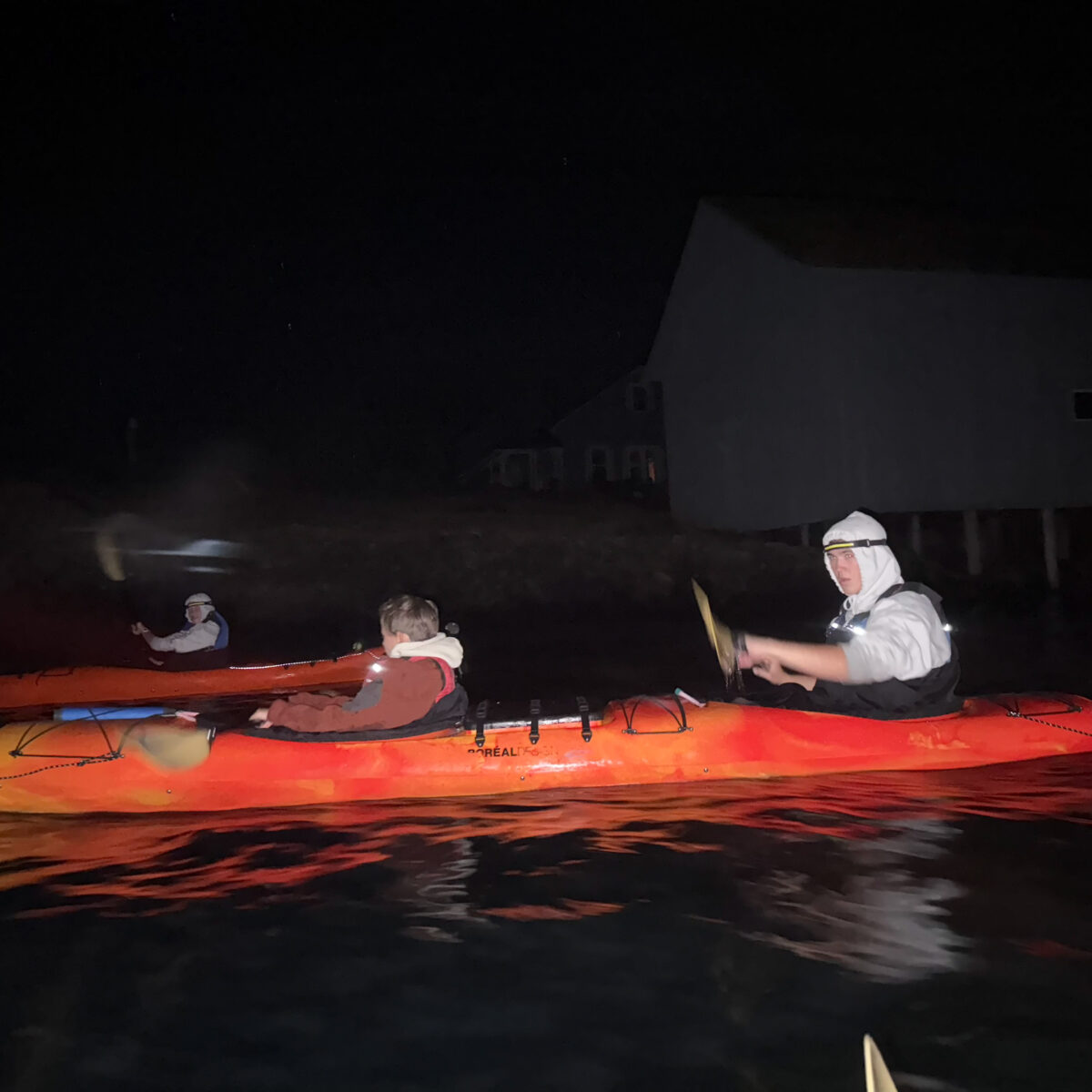 Three people in orange kayaks paddle on dark water at night near Friday Harbor Kayaking, illuminated by a camera flash. Two houses are visible in the background.