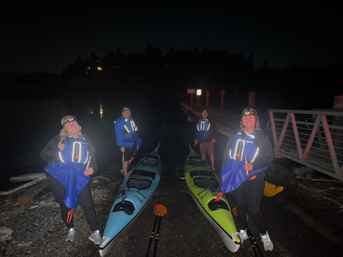 Four people with headlamps stand next to two kayaks on a rocky shore at night, near a dock and calm water—ready for a Friday Harbor Kayaking adventure under the stars.