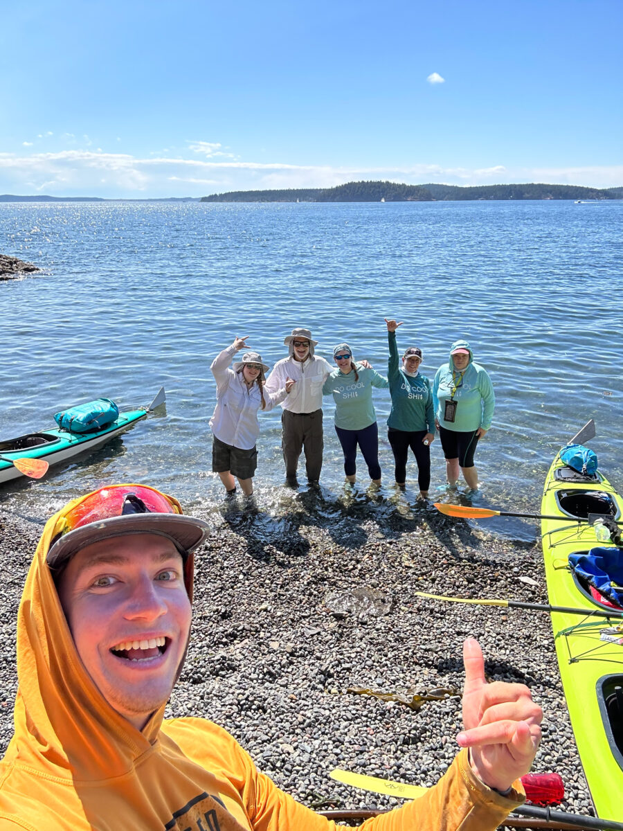 Washington Kayaking Guide to the Best Places, Wildlife, and Guided Tours 1 A group of five people poses on a rocky shoreline with kayaks beside them during a Washington Kayaking trip, while one person in the foreground gives a thumbs up and smiles at the camera. The water and island are visible in the background.