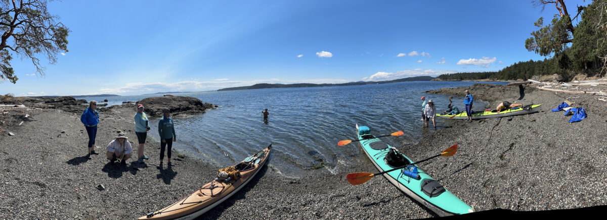 People stand and prepare kayaks on a rocky beach by the sea, with clear skies and distant islands visible across the water—an ideal scene for those seeking memorable Kayaking Tours in Seattle.