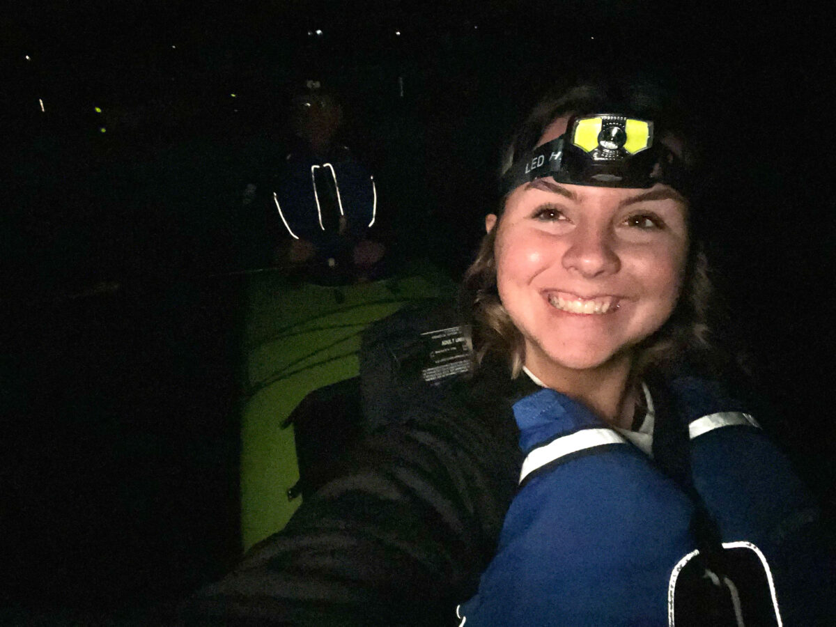 A person wearing a headlamp and life jacket smiles for a selfie while kayaking at night with Friday Harbor Kayaking; another paddler is faintly visible in the background.