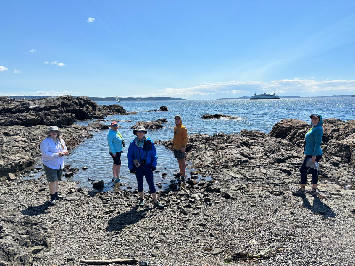 Five people stand on a rocky shoreline under a clear sky, with a San Juan Islands Boat Tours ferry visible in the distance on the water.