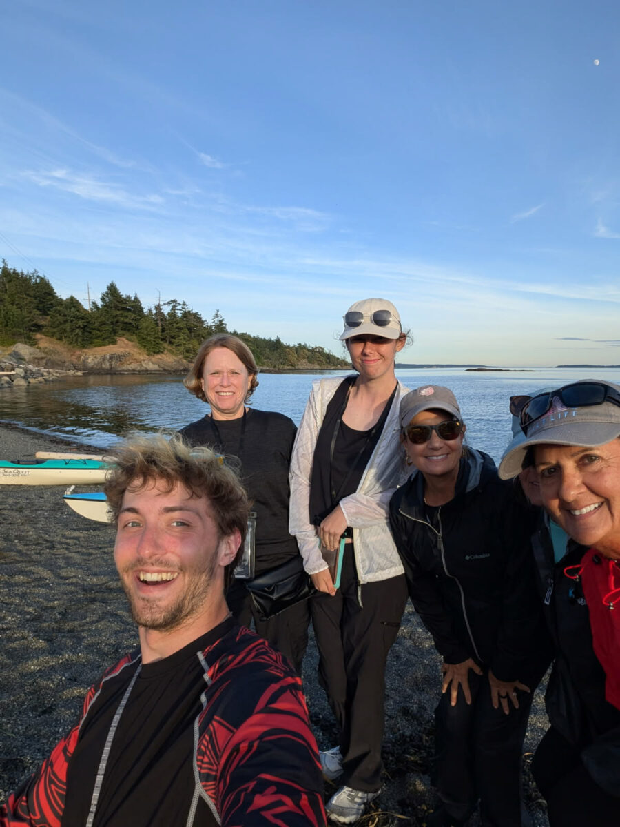 Washington Kayaking Guide to the Best Places, Wildlife, and Guided Tours 2 Five people pose and smile on a rocky beach near water and paddleboards, enjoying a sunny day of Washington kayaking with trees and a clear blue sky in the background.