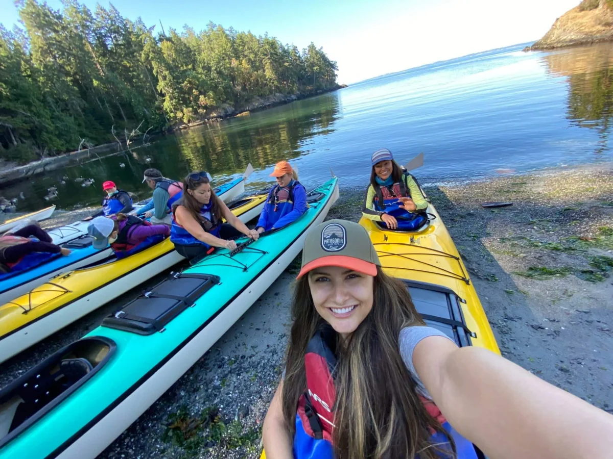A group of people in life jackets prepare for kayak tours on calm water near a forested shoreline; a woman in front takes a selfie.