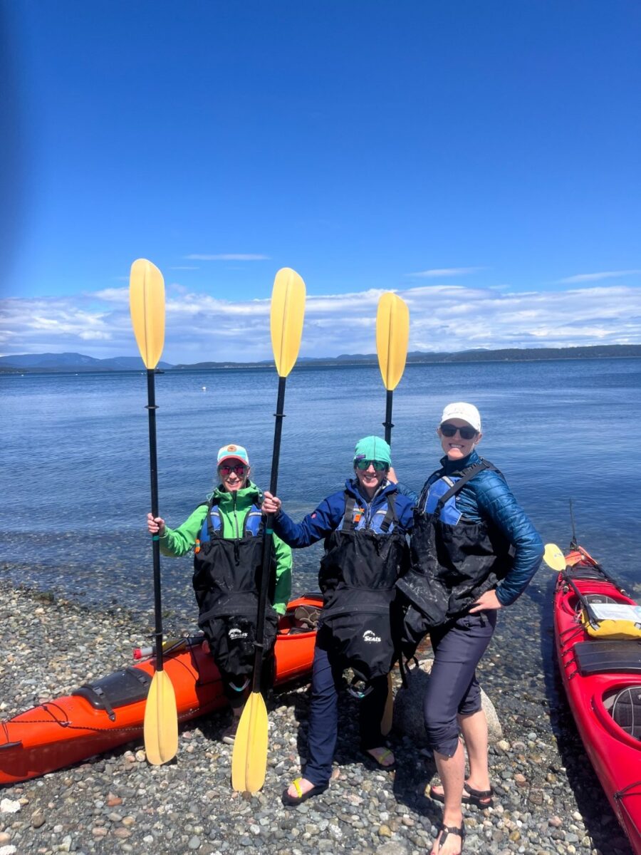 This Week on the Water: Orcas, Bioluminescence & a Full Slate of Day Trips (June 29 – July 5, 2026) 2 Three guided sea kayak paddlers on a rocky Friday Harbor beach with orange and red sea kayaks, San Juan Islands, Washington — Sea Quest Kayak Tours